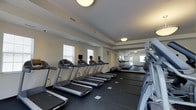 This single-level fitness center is designed to provide a functional and comfortable workout environment. The space is laid out with a row of treadmills equipped with individual display monitors along one side, allowing users to track their workouts while exercising. To the right, strength and cardio equipment adds variety to the available workout options, while exercise balls are neatly stored toward the back of the room.

The interior features neutral-toned walls and dark rubberized flooring, which is practical for high-traffic exercise use. Flush-mounted ceiling lights provide consistent, even illumination throughout. A large mirrored wall at the far end of the room visually expands the space and allows exercisers to monitor their form. Windows fitted with blinds allow natural light to filter in while maintaining privacy.

The overall layout is clean and organized, with sufficient spacing between equipment to allow for comfortable movement. The mirrored rear wall also reflects a set of glass doors, suggesting access to an adjacent area or exterior, contributing to the open and airy ambiance of the fitness center.