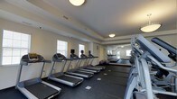 This single-level fitness center is designed to provide a functional and comfortable workout environment. The space is laid out with a row of treadmills equipped with individual display monitors along one side, allowing users to track their workouts while exercising. To the right, strength and cardio equipment adds variety to the available workout options, while exercise balls are neatly stored toward the back of the room.

The interior features neutral-toned walls and dark rubberized flooring, which is practical for high-traffic exercise use. Flush-mounted ceiling lights provide consistent, even illumination throughout. A large mirrored wall at the far end of the room visually expands the space and allows exercisers to monitor their form. Windows fitted with blinds allow natural light to filter in while maintaining privacy.

The overall layout is clean and organized, with sufficient spacing between equipment to allow for comfortable movement. The mirrored rear wall also reflects a set of glass doors, suggesting access to an adjacent area or exterior, contributing to the open and airy ambiance of the fitness center.