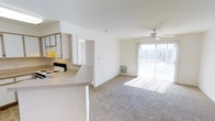 This single-floor residence offers three bedrooms and two bathrooms within a thoughtfully arranged open layout. The living area is carpeted and bathed in natural light streaming through large sliding glass doors that lead to an outdoor area, creating a bright and welcoming atmosphere. A ceiling fan overhead adds both function and style to the main living space, while a wall vent ensures year-round climate comfort.

The kitchen is positioned adjacent to the living area in a semi-open configuration, featuring white cabinetry, an electric range, and a countertop peninsula that provides a natural division between the cooking and living spaces. The light-toned cabinetry and white appliances contribute to a clean, cohesive aesthetic throughout.

A doorway visible from the main living area leads to the private quarters of the home, offering easy access to the bedrooms and bathrooms. The overall design emphasizes practicality and flow, with each area connecting seamlessly to the next. The abundance of natural light, open-plan arrangement, and neutral color palette create a versatile and move-in ready environment suitable for a range of lifestyles and furnishing preferences.
