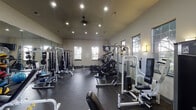 This fitness center in Jersey Village, TX, offers a comprehensively outfitted workout facility designed to support a wide range of exercise routines. The single-floor space features soaring vaulted ceilings that lend an open, airy feel, complemented by recessed lighting, ceiling fans, and wall-mounted sconces that ensure the space is well-lit throughout the day and evening. Multiple large windows line the walls, flooding the interior with natural light and providing views of the surrounding greenery outdoors.

The flooring throughout is durable dark rubber, ideal for high-impact training and heavy equipment use. The strength training area is generously appointed with cable machines, weight stack systems, a squat rack, adjustable benches, a full dumbbell rack, and an assortment of medicine balls and stability balls. Floor-to-ceiling mirrors along one wall enhance the sense of space and allow members to monitor their form during workouts.

The cardio section features stair climbers, a stationary cycle, and treadmills, all positioned near the windows to take advantage of the natural light and outdoor views. Wall-mounted televisions are also present for entertainment during workouts. Overall, the fitness center delivers a functional, well-maintained environment suitable for both strength and cardiovascular training.