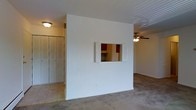 This single-story space is characterized by a functional and straightforward layout. A prominent interior wall serves as the dividing element between the living area and the kitchen, featuring a pass-through window fitted with white bi-fold shutters. This design detail allows for visual connectivity and convenience between the two spaces while maintaining defined zones. Through the pass-through, dark reddish-brown cabinetry and kitchen appliances are visible, hinting at a compact but practical kitchen area. The living area is finished with wall-to-wall carpeting in a neutral tone, contributing to a warm underfoot feel. A ceiling fan with a light fixture is mounted overhead, providing both air circulation and ambient lighting. To the left, a separate area is accessible via a flush transition from carpet to smooth flooring, where a set of bifold closet doors provides generous storage. The walls throughout are painted in a clean, neutral white, and flush-mount lighting fixtures illuminate the space. Overall, the layout prioritizes practicality, with thoughtful details like the pass-through window adding a degree of openness to the interior design.