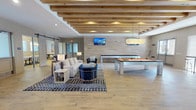 This well-appointed clubhouse is designed to offer a welcoming and versatile social environment. Exposed natural wood ceiling beams are a defining architectural feature throughout the space, adding warmth and a modern-rustic aesthetic that contrasts beautifully with the clean, neutral-toned walls and light wood-look plank flooring underfoot.

The main gathering area is generously sized, accommodating multiple distinct lounge zones furnished with contemporary sofas, armchairs, and patterned area rugs. A wall-mounted flat-screen TV anchors one end of the room, while a linear fireplace with white brick surround adds ambiance to a secondary seating area equipped with a mounted TV, built-in shelving, and a striking multi-arm chandelier. Large windows with plantation shutters allow ample natural light to fill the space.

The game area features a full-size billiards table alongside a shuffleboard table, making it ideal for casual recreation. A sliding barn door provides separation from a corridor leading to additional spaces. The kitchenette is equipped with a full-size stainless steel refrigerator, ample cabinetry, and a dedicated coffee station with counter seating nearby, ensuring residents have convenient access to refreshments throughout their visit.