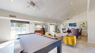 Located at 5220 W Northern Ave in Glendale, Arizona, this well-appointed recreation and lounge space offers a versatile, open-plan layout designed for both leisure and socializing. The room features vaulted ceilings with ceiling fans and recessed lighting that keep the atmosphere bright and airy throughout.

A centerpiece billiards table anchors the main area, positioned beneath soaring ceilings that lend a generous sense of volume to the space. Large sliding glass doors and floor-to-ceiling windows line the exterior walls, flooding the interior with abundant natural light and providing seamless views and direct access to an outdoor patio and pool area.

The lounge zone is thoughtfully furnished with upholstered wingback chairs, ottomans, and a central cocktail table arranged around a wall-mounted flat-screen television, flanked by contemporary abstract artwork. A rustic wooden console serves as a coffee station, adding a warm, functional touch to the seating area.

A compact kitchenette along one wall includes cabinetry, a stainless steel refrigerator, and a microwave, supporting the space's amenity-focused design. Light-toned tile flooring runs throughout, complementing the crisp white walls and modern, eclectic furnishings. The overall ambiance is clean, contemporary, and inviting — ideal as a community amenity space.