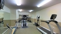 This fitness center offers a practical and well-organized space dedicated to health and exercise. The room features a textured ceiling with bright fluorescent lighting that ensures the area is well-lit throughout. The flooring is carpeted, providing a comfortable surface underfoot, while the walls are finished in a two-tone paint scheme with a warm beige lower half and a lighter upper half, creating a clean and neutral aesthetic. A large wall-to-wall mirror along the back wall visually expands the space and allows users to monitor their form during workouts. The facility is equipped with an impressive range of exercise machines, including a treadmill, elliptical trainer, upright exercise bike, and a multi-function cable and weight machine, catering to both cardiovascular and strength training needs. Additional equipment is visible in the mirrored reflection, suggesting further options for a varied fitness routine. The layout is thoughtfully arranged to allow comfortable movement between stations. The space presents a functional, no-frills environment ideal for residents or members seeking convenient access to essential fitness amenities.