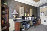 Located in Woodstock, Georgia, this single-floor business center offers a well-appointed space designed to blend functionality with traditional elegance. The room features distinctive dark patterned wallpaper paired with cream-painted wainscoting, creating a sophisticated, classic aesthetic throughout.

A dedicated computer workstation area lines one wall, equipped with multiple desktop computers, a long granite-topped desk, and ergonomic task chairs, providing a productive environment for work or research. Framed artwork and decorative wall sconces accent this section, adding a refined touch to the practical workspace.

On the opposite side, a cozy seating nook features a cushioned armchair and a small decorative side table, offering a comfortable spot for reading or relaxing. A brochure rack nearby provides easy access to local information and area guides.

The room benefits from a large window fitted with blinds, overlooking an outdoor pool area and allowing natural light to fill the space. A graceful barrel-vaulted ceiling with coffered detailing adds architectural interest and a sense of openness overhead.

A glass-paned French door connects the business center to adjacent areas, enhancing the flow between spaces. Patterned carpeting ties the room together, complementing the overall warm and welcoming ambiance of this well-equipped facility.