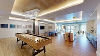 This single-floor community lounge is a thoughtfully designed, multi-functional gathering space that blends recreational amenities with relaxed social areas. The open-plan layout is anchored by warm wood-look flooring throughout, complemented by a mix of natural and ambient lighting, including a distinctive coffered ceiling with warm LED cove lighting and a modern geometric chandelier.

The main activity zone features a full-length shuffleboard table as a standout focal point, positioned beneath the chandelier for a polished, game-room aesthetic. Adjacent to this area, a comfortable lounge section offers plush seating, a wall-mounted flat-screen television, and decorative wall art, creating an inviting space for informal gatherings.

A built-in kitchenette with dark cabinetry, a sink, and counter seating serves as a practical hub for social events. Nearby, a bar-height island provides additional seating. A quieter nook features a gallery wall of framed artwork, high-top seating, and a mounted display, ideal for smaller meetings or focused activities.

The secondary lounge area showcases a mid-century modern aesthetic, with a neutral sofa, a rocking chair, a bold patterned area rug, and built-in open shelving. Large windows allow natural light to filter in, while glass entry doors connect the space to the outdoors, enhancing the overall sense of openness.