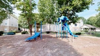 This outdoor playground and recreational area offers a variety of amenities designed for active enjoyment in a well-maintained community setting. The centerpiece of the space is a multi-component play structure finished in blue and white, featuring an elevated platform with railed walkways, a spiral climbing element, and multiple slides for children of varying ages. A separate swing set is positioned nearby on a open dirt play surface, providing additional recreation options.

Towering mature trees provide generous natural shade throughout the playground area, creating a comfortable outdoor environment during warmer months. The grounds are landscaped with low shrubs and green grass that frame the space attractively.

Adjacent to the playground is a large outdoor swimming pool with a spacious concrete deck, furnished with lounge chairs and shaded by umbrella-equipped seating areas — ideal for relaxation. Stone columns with a stacked natural rock finish accent the pool area, lending a refined architectural touch that is echoed in the exterior detailing of the surrounding multi-story residential building. The building itself features a combination of neutral lap siding and natural stone cladding at its base.

A covered outdoor patio with wood furniture and a grill provides an inviting communal gathering spot, completing this well-rounded outdoor recreational amenity space.