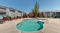 Located at 4407 Oakhollow Dr in Sacramento, California, this outdoor community amenity area serves as a central gathering space within a multi-building residential complex. The centerpiece is a generously sized freeform swimming pool with a smooth, curved design, complemented by an attached spa for relaxation. The expansive concrete pool deck surrounds the water on all sides, providing ample space for sunbathing and leisure, and is furnished with multiple bold red lounge chairs arranged along the poolside. The area is enclosed by a black iron fence with gated entry and exit points, ensuring privacy and security for residents. A wooden pergola-style structure near one end of the deck adds a shaded area for additional outdoor use. Adjacent to the pool is a single-story clubhouse building with board-and-batten siding, large windows, and double glass doors that offer a direct view of the pool area. Mature trees surrounding the complex provide natural shade and a touch of greenery against the clear Sacramento sky. The multi-story residential buildings visible in the background feature balconies and an exterior staircase design typical of established community complexes. Overall, the space offers a well-maintained, functional outdoor retreat suited for relaxation and community enjoyment.