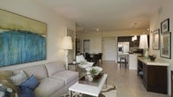 This single-floor residence offers an open-concept layout that connects the living, dining, and kitchen areas in one fluid, well-lit space. The living area is anchored by a plush gray sofa, a sleek white coffee table, and a patterned area rug, complemented by a bold abstract artwork that adds a pop of color to the neutral walls. A contemporary floor lamp provides warm ambient lighting, enhancing the inviting atmosphere.

Adjacent to the living area, the dining space accommodates a dark-toned table and chairs, creating a subtle contrast against the light tile flooring that runs throughout the unit. A white paneled door leads to an additional room, offering functional versatility.

The kitchen is clearly visible from the main living area, featuring dark cabinetry, stainless steel appliances including a refrigerator with an ice dispenser, and a peninsula counter with bar-height seating. Pendant lighting above the kitchen adds a refined touch. The light-toned tile flooring unifies all areas, while smooth, neutral-colored walls and track lighting contribute to the clean, modern aesthetic of the space.
