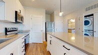 Located at 140 E Monument Ave in Dayton's historic Monument Avenue corridor, this single-floor unit presents a clean, modern aesthetic throughout. The kitchen serves as a central highlight, featuring white shaker-style cabinetry with matte black hardware, gray countertops, and a full suite of stainless steel appliances including a range, microwave, dishwasher, and refrigerator. A substantial center island with pendant lighting above provides additional prep and seating space, while light wood-tone flooring ties the area together and extends into the main living space.

The open layout connects the kitchen to a generously sized living area, which benefits from large windows that invite natural light and offer views of surrounding greenery. The bedrooms are finished with plush neutral carpeting and feature bright windows, creating a comfortable and quiet retreat. One bedroom includes direct access to a well-appointed en-suite bathroom.

The bathroom is outfitted with a dual-sink vanity, white cabinetry with matching matte black hardware, a large mirror, gray countertops, and a tub-shower combination. Wood-look flooring carries through the bathroom, maintaining a cohesive design language. The overall palette of whites, grays, and warm wood tones gives the unit a fresh, contemporary feel throughout.