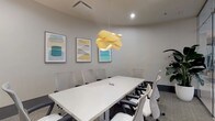 This single-floor conference space is thoughtfully designed to balance professional functionality with refined aesthetic appeal. The centerpiece of the room is a large, sleek white conference table surrounded by ergonomic white and gray task chairs, comfortably accommodating multiple attendees. A striking sculptural pendant light in warm yellow tones hangs above the table, serving as an eye-catching focal point that adds warmth to the otherwise neutral palette of soft gray walls and textured carpet flooring.

One wall features a dark credenza topped with decorative accents, including a gold starburst sculpture and table lamps, alongside a wall-mounted flat-screen television — making the room well-equipped for presentations and collaborative work. A series of colorful abstract artworks in muted teal, yellow, and gray tones are displayed along the walls, lending a creative energy to the space.

Glass partition walls with sliding barn-style hardware define the room's boundary while maintaining a visual connection to the adjoining common area, allowing natural light to filter in and creating an open, airy feel. A large potted tropical plant positioned in the corner introduces a refreshing organic element. The overall ambiance is polished, modern, and conducive to focused professional gatherings.