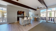 Located on Mission Blvd in Hayward, California, this single-floor space presents a thoughtfully designed open-concept layout that seamlessly blends functional and aesthetic elements throughout. Striking dark wood exposed beams run across the vaulted ceiling, lending architectural character and a sense of warmth to the interior.

The kitchen is accessed through a distinctive arched opening and features rich dark wood cabinetry, dark stone countertops, a geometric mosaic tile backsplash, and stainless steel appliances including a built-in microwave. A central island with decorative patchwork tile detailing and dark countertop serves as a natural gathering point, surrounded by modern white bar stools with pedestal bases. Light wood-style flooring flows through the kitchen and dining areas.

The expansive living area is carpeted and anchored by a floor-to-ceiling stacked stone fireplace with a wall-mounted television. Generous sliding glass doors and large windows flood the space with natural light and open to a balcony with outdoor views. Ceiling fans are installed throughout, enhancing comfort. A cozy seating nook with tufted accent chairs and wall-mounted planters adds a charming, personalized touch. The overall ambiance is modern yet inviting, with a cohesive neutral palette accented by organic textures.