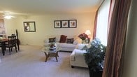 This two-floor residence offers a comfortable and well-designed living environment. The living room serves as a central gathering space, featuring soft wall-to-wall carpeting that adds warmth underfoot. Large windows dressed with elegant floor-length drapes in rich red tones allow generous natural light to fill the room, creating a bright and inviting atmosphere. The space comfortably accommodates a white upholstered sofa and matching armchair, arranged around a circular glass-topped coffee table with a wooden base. Decorative touches, including a trio of framed artwork displayed on the wall and lush potted plants, contribute to the room's polished yet relaxed aesthetic. A floor lamp provides warm ambient lighting, complementing the natural light from the windows. The neutral, cream-toned walls and light carpeting create a versatile backdrop that enhances the sense of openness. Spread across two floors, this residence provides a practical and spacious layout suited for everyday living, with the ground level anchored by this well-appointed living area that sets a welcoming tone throughout the home.