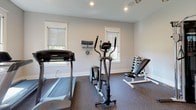 Located at 5 Patriots Place in Millbury, Massachusetts, this single-floor fitness space is thoughtfully designed to support a comprehensive workout experience. The room is finished with neutral gray walls and durable rubber flooring, providing a clean, functional environment suited for a range of exercise activities.

The space is outfitted with an impressive array of equipment, including a treadmill, elliptical trainer, stationary spin bike, a multi-function cable machine, an adjustable weight bench, a dumbbell rack, and a stability ball. Wall-mounted accessories add further utility, keeping the area organized and efficient.

Natural light filters in through multiple windows fitted with roller shades, creating a bright and energizing atmosphere. Recessed ceiling lighting and a ceiling fan supplement illumination and airflow throughout the room. A wall-mounted flat-screen television adds an entertainment element for those working out.

A standout architectural detail is the set of white-trimmed French doors with divided glass panes, which open to an adjacent space featuring hardwood flooring, a pendant lantern fixture, and what appears to be a lounge or refreshment area. A fire alarm system is also visibly present, reflecting attention to safety standards throughout the space.