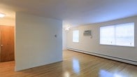 This single-floor space offers a clean and functional layout with a welcoming, understated aesthetic. The main living area is open and generously sized, showcasing gleaming hardwood floors that reflect natural light pouring in through multiple windows. The bright, neutral white walls contribute to an airy and expansive feel throughout the room. A ceiling fan provides overhead comfort and adds a subtle decorative touch to the space. Climate control is well-equipped, with a wall-mounted air conditioning unit and a baseboard heating system ensuring year-round comfort. The windows, framed simply and efficiently, allow ample daylight to fill the room, enhancing its open character. The layout is straightforward and versatile, offering flexibility for various furniture arrangements and uses. Overall, the space presents a practical and light-filled environment with durable finishes and essential amenities in place.
