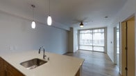 This single-floor space features an open-concept layout that seamlessly connects the kitchen and living areas. The kitchen is equipped with a stainless steel sink set into a light-toned countertop, complemented by warm wood cabinetry and two pendant lights that hang above, providing focused task lighting with a modern aesthetic. The living area extends naturally from the kitchen, finished with smooth hardwood-style flooring that runs throughout the space, adding warmth and continuity. A ceiling fan is centrally positioned in the living area, offering both practical airflow and a subtle design element. Floor-to-ceiling windows along the far wall allow an abundance of natural light to fill the interior, creating a bright and airy atmosphere while offering views of the surrounding urban environment. The walls are finished in a clean, neutral tone that enhances the sense of openness. The overall design is contemporary and minimal, with a cohesive palette of light tones and clean lines that give the space a functional yet polished character.