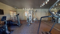 This single-floor fitness room offers a focused exercise environment designed to accommodate a variety of workout routines. The space is outfitted with several pieces of cardio equipment, including elliptical trainers, a stair climber, and a stationary recumbent bike, providing options for cardiovascular conditioning. A multi-station cable weight machine serves as the centerpiece for strength training, complemented by an upright rowing or climbing apparatus nearby.

The room features patterned, low-pile carpeting throughout, lending a practical yet finished appearance to the floor. Track lighting runs along the ceiling, directing focused illumination across the equipment. A full-length mirrored wall spans one side of the room, visually expanding the space and allowing users to monitor their form during exercise. The darker-toned walls and ceiling contribute to a contained, utilitarian atmosphere.

Natural light enters through shuttered windows on one wall and a set of double glass-panel doors on the opposite end, which also serve as an exit. Posted signage near the entrance outlines facility rules and guidelines. Overall, the fitness room is efficiently organized to maximize usable floor space while providing a functional range of equipment for both cardio and strength-based training.