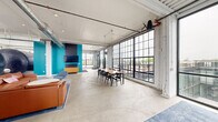 This single-floor space delivers a compelling industrial aesthetic rooted in adaptive reuse design. The open main area is defined by polished concrete floors, exposed ductwork running across white-painted ceilings, and an expansive bank of black steel-framed, floor-to-ceiling windows that flood the interior with natural light and offer views of the surrounding rooftop terrace and cityscape beyond. Oversized repurposed industrial tanks serve as dramatic architectural centerpieces — one has been ingeniously converted into a semi-private lounge nook, finished with warm brass-toned metal, built-in wood bench seating, vivid magenta cushions, and pendant lighting, creating an intimate retreat within the larger space. The main lounge area features a generous sectional leather sofa arrangement paired with a round accent chair and coffee table, anchored by a blue area rug. A wall-mounted TV is set against a bold teal accent wall. A separate dining zone provides a long communal table with ample seating. Access to the outdoor terrace is provided through glass doors. A fully tiled, ADA-accessible restroom with a frosted-glass teal door completes the layout. The overall ambiance is energetic yet functional, balancing industrial history with modern communal living.