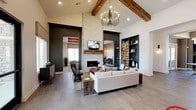 This single-story clubhouse showcases a striking blend of rustic and contemporary design elements throughout its well-appointed spaces. The main lounge commands attention with soaring vaulted ceilings supported by thick, exposed wood beams and anchored by a floor-to-ceiling herringbone-tiled fireplace with a live-edge wood mantel and wall-mounted television. A sculptural chandelier hangs overhead, while large-format tile flooring, a plush neutral sectional, and built-in shelving displaying stacked log rounds complete the inviting atmosphere.

Adjacent to the lounge, a dining area features vibrant red lattice-back chairs paired with dark round tables beneath the same impressive vaulted ceiling, positioned near a kitchen bar with bold red glazed tile walls, dark cabinetry, white countertops, and a double sink — ideal for entertaining.

A dedicated lounge nook offers a distinctive coffered ceiling painted in deep orange, textured accent walls, metallic stump-style tables, and sculptural seating alongside a built-in workstation with computer access. Natural light filters throughout via multiple windows with exterior views, tying together this energetic, design-forward communal space.