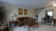 This two-floor home offers a traditionally styled living space with a warm and inviting ambiance. The main living room is finished with plush wall-to-wall carpeting and neutral-toned walls, creating a clean and understated backdrop. A classic sofa with rich, patterned upholstery serves as the focal point of the room, accompanied by wood-accented side tables and a decorative coffee table with turned legs and carved detailing. Framed botanical artwork adorns the walls, adding a touch of character to the space. A pair of traditional table lamps provides warm ambient lighting throughout the room. A staircase with a white banister is visible toward the back of the room, connecting the main level to the upper floor. The layout of the living area suggests a comfortable flow between spaces, with room for additional seating or accent pieces. The overall design aesthetic leans toward a classic, traditional style, with consistent use of wood tones and soft textiles that contribute to a lived-in, homey feel throughout the space.