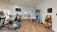 This single-level fitness room is thoughtfully designed to support a full range of workout routines. The space is finished with warm hardwood-style flooring throughout, contributing to a clean and inviting atmosphere. White walls adorned with bold motivational typography — featuring words such as "commitment," "focus," "strength," and "dedication" — create an energizing environment that reinforces a fitness-forward mindset.

The room is well-lit by multiple large windows fitted with white blinds, allowing generous natural light to fill the space. Track lighting on the ceiling provides additional illumination. A wall-mounted flat-screen TV is positioned for easy viewing during workouts.

The equipment selection is comprehensive, including a treadmill, elliptical trainer, stationary recumbent bike, and a multi-functional cable resistance machine equipped with adjustable pulleys and resistance bands for versatile strength training. A set of French doors connects the fitness room to an adjacent interior area, enhancing the sense of openness and flow. Overall, the space offers a bright, functional, and motivating environment well-suited for dedicated fitness use.