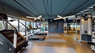 This fitness center is designed with both functionality and aesthetics in mind, offering a comprehensive workout environment across a single level. The space is divided into distinct zones, beginning with a spacious cardio area featuring multiple treadmills, stair climbers, and elliptical machines positioned along expansive floor-to-ceiling windows that frame outdoor city views, flooding the area with natural light.

The flooring throughout the main cardio zone features a durable speckled rubber surface with contrasting tan and grey striped inlays, providing both visual interest and practical cushioning underfoot. The industrial-style ceiling is finished in a deep teal-blue tone with exposed structural elements and linear pendant lighting that adds a modern, polished atmosphere.

A dedicated strength training area is separated by glass-paneled partitions with frosted geometric patterns, housing cable machines, a rowing machine, and weight benches. Wall-mounted mirrors enhance the sense of space in this section. Mounted televisions are positioned throughout the facility for entertainment during workouts. The overall design balances an energetic, contemporary aesthetic with practical, high-quality equipment and thoughtful spatial organization.