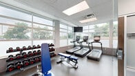 Located at 2521 Kingston Pike in Knoxville, Tennessee, this fitness facility offers a well-appointed, single-floor exercise space designed to support a comprehensive workout experience. The layout is bright and open, benefiting from an expansive wall of large windows that flood the interior with natural light and provide views of the surrounding greenery.

The main exercise area is equipped with a wide array of fitness equipment, including treadmills, ellipticals, a stair climber, and a cable machine, catering to both cardio and strength training needs. A full rack of dumbbells lines one wall alongside a weight bench, while medicine balls and kettlebells are neatly organized near floor-to-ceiling mirrors that enhance the sense of space throughout the room. The durable speckled flooring throughout the facility is both functional and easy to maintain.

A dedicated relaxation area features a massage lounger and wall-mounted television, offering members a space to recover after their workout. The facility also includes an additional room framed by a frosted glass door, providing a private or multifunctional area. The drop ceiling with recessed panel lighting ensures consistent, even illumination throughout. Overall, this facility delivers a clean, modern, and fully functional environment for fitness and wellness.