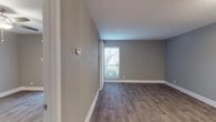 This single-floor space presents a clean, modern aesthetic throughout, unified by durable wood-look flooring and neutral gray wall tones complemented by crisp white baseboards. The layout, visible in the overhead floor plan, includes a living area, kitchen, bedroom, and bathroom arranged for functional flow.

The living area is an open, well-proportioned room featuring a window with horizontal blinds that admits natural light and outdoor views. A thermostat is conveniently mounted on the wall, and the space connects directly to the kitchen and interior hallway.

The kitchen is thoughtfully updated with white shaker-style cabinetry, light marble-look countertops, a white subway tile backsplash, and a full suite of stainless steel appliances including a refrigerator, dishwasher, and range. A window above the sink provides additional natural light.

The bedroom features a ceiling fan with integrated lighting and a sliding door with vertical blinds, offering access or views to an outdoor area. An additional room provides flexible use as a second bedroom or office, with a flush-mount ceiling fixture.

The bathroom includes a full tub and shower combination lined with large-format marble-style tile in soft neutral tones, fitted with brushed nickel fixtures for a polished finish.
