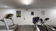 Located at 500 Rolling Hills Pl in Lancaster, Texas, this fitness center offers a well-organized and fully equipped exercise space designed to support a variety of workout routines. The room features durable rubber flooring throughout, providing a safe and practical surface for both cardio and strength training activities. Large wall-mounted mirrors line multiple walls, visually expanding the space and allowing users to monitor their form during exercise. Natural light filters in through windows fitted with blinds, complementing the recessed and overhead fluorescent lighting to keep the space bright and functional. The cardio equipment selection includes multiple treadmills, an elliptical trainer, a stair climber, and both upright and recumbent stationary bikes, catering to a range of fitness preferences and skill levels. A set of free weights and a weight bench are also available for strength training. Wall-mounted flat-screen televisions provide entertainment during workouts, while motivational posters and a core exercise reference chart add an instructional and energizing ambiance. The space is clearly marked with posted gym rules and an accessible exit, ensuring a safe and organized environment. Overall, this fitness center delivers a comprehensive and practical workout facility in a clean, no-frills setting.