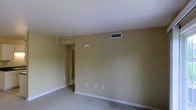 This single-story space presents a straightforward and functional layout. The main room is finished with wall-to-wall carpeting in a neutral tone, providing a clean and understated foundation. The walls are painted in a warm beige, contributing to a simple, uniform aesthetic throughout. The ceiling is smooth and white, featuring multiple HVAC vents that indicate a central air system, ensuring climate control throughout the space. A small thermostat or utility panel is mounted on the wall, adding to the practical nature of the room. Multiple electrical outlets are evenly distributed along the baseboards, offering convenient access for various needs. White baseboards provide a clean contrast against the carpeted floor and beige walls. A hallway visible from the main room leads to additional areas of the unit, suggesting a connected and accessible floor plan. Natural light appears limited based on the current view, though the open layout helps maintain a sense of openness. Overall, the space is move-in ready with a clean, no-frills design suited for practical, everyday living.