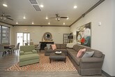 This single-floor community clubroom offers a well-appointed gathering space designed for both relaxation and casual socializing. The layout is thoughtfully divided into two primary zones: a comfortable lounge area and a sunlit dining section, each with its own distinct character while maintaining a cohesive aesthetic throughout.

The lounge area anchors the space with a generously sized charcoal sectional sofa, accented by colorful throw pillows and set atop a bold orange-and-white patterned area rug. A large-scale abstract painting in vivid blues, yellows, and reds commands the wall behind the seating, adding an energetic focal point. Recessed lighting and a ceiling fan provide both illumination and comfort, while dark crown molding adds architectural definition.

Adjacent to the lounge, the dining area features multiple round tables with upholstered chairs, ideal for group gatherings. Floor-to-ceiling windows line the perimeter of this section, flooding the space with natural light and providing unobstructed views of the outdoor playground and surrounding residential grounds. Wood-look plank flooring runs throughout, unifying both zones seamlessly.

Decorative accents, including an oversized wall clock and curated artwork, contribute to the space's warm, welcoming ambiance.