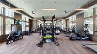 Located at 900 Corby Lane in Charleston, SC, this fitness center offers a well-appointed, single-floor exercise space designed to support a comprehensive range of workout routines. The room features elevated ceilings with a tiered tray design, pendant linear lighting fixtures, and ceiling fans that promote air circulation throughout the facility. Large, multi-pane windows line the perimeter walls, flooding the interior with generous natural light and providing views of the surrounding greenery. The flooring consists of durable wood-look tile in a warm neutral tone, lending a modern yet approachable aesthetic. Full-length mirrors span one entire wall, enhancing the sense of openness while providing practical feedback during workouts. The equipment layout is thoughtfully organized, with a dedicated cardio zone featuring treadmills, elliptical trainers, and a stair climber positioned near the windows. A central multi-functional rack serves as the focal point of the strength training area, surrounded by a full dumbbell rack, kettlebells, battle ropes, stability balls, a BOSU balance trainer, and weight benches. Selectorized strength machines and a cable system round out the offerings. A water fountain is also available on-site. The neutral taupe wall tones and clean architectural lines create a focused, motivating environment suited for all fitness levels.