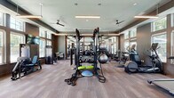 Located at 900 Corby Lane in Charleston, SC, this fitness center offers a well-appointed, single-floor exercise space designed to support a comprehensive range of workout routines. The room features elevated ceilings with a tiered tray design, pendant linear lighting fixtures, and ceiling fans that promote air circulation throughout the facility. Large, multi-pane windows line the perimeter walls, flooding the interior with generous natural light and providing views of the surrounding greenery. The flooring consists of durable wood-look tile in a warm neutral tone, lending a modern yet approachable aesthetic. Full-length mirrors span one entire wall, enhancing the sense of openness while providing practical feedback during workouts. The equipment layout is thoughtfully organized, with a dedicated cardio zone featuring treadmills, elliptical trainers, and a stair climber positioned near the windows. A central multi-functional rack serves as the focal point of the strength training area, surrounded by a full dumbbell rack, kettlebells, battle ropes, stability balls, a BOSU balance trainer, and weight benches. Selectorized strength machines and a cable system round out the offerings. A water fountain is also available on-site. The neutral taupe wall tones and clean architectural lines create a focused, motivating environment suited for all fitness levels.