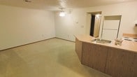 This single-floor space features an open-concept layout that combines the living area and kitchen into one cohesive, easy-to-navigate floor plan. The living area is generously sized, finished with wall-to-wall carpeting in a neutral tone that lends warmth to the space. A ceiling fan is centrally mounted, providing both lighting and air circulation. The kitchen is defined by a wood-paneled peninsula that extends into the living area, topped with a built-in sink and offering a natural division between the two spaces. A white refrigerator is visible alongside the peninsula, completing the kitchen's functional setup. The walls are finished in a clean, neutral white throughout, complemented by standard baseboards that frame the room neatly. A doorway visible toward the rear of the space leads to an additional room, suggesting a separate bedroom or utility area beyond. The textured ceiling adds a subtle design detail consistent with the overall practical aesthetic. The layout is straightforward and efficient, with ample open floor space that provides flexibility for a variety of furniture arrangements. Overall, the space offers a clean, functional environment with a simple, understated character.