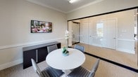 This single-floor meeting room showcases a refined and professional design, defined by its striking floor-to-ceiling glass partition walls framed in black, which provide both visual openness and a clear boundary from the adjacent workspace. The glass walls allow views into the broader office environment while maintaining an intimate and focused atmosphere within the room itself. Crown molding along the upper perimeter adds a touch of architectural elegance to the neutral beige walls, which are complemented by a chair rail detail that runs the length of the room. Textured carpet flooring in warm tan tones adds comfort underfoot. A round white pedestal table serves as the centerpiece, surrounded by contemporary gray upholstered chairs, creating a welcoming space ideal for small meetings or collaborative discussions. A wall-mounted flat-screen display on one side supports presentations and video conferencing, while a dark wood credenza offers functional storage. Natural light filters in generously through tall windows that frame greenery and outdoor views, enhancing the room's bright and pleasant ambiance. A framed abstract artwork and a potted white orchid add refined decorative touches, completing the polished, professional character of this meeting space.