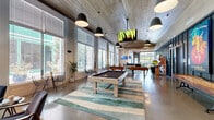 Located at 345 Heatherden Ave in Fayetteville, Georgia, this single-floor community amenity center offers a generously sized, open-plan layout designed for both recreation and productivity. The space showcases a compelling blend of modern industrial and contemporary design, highlighted by soaring ceilings clad in warm wood planking, polished concrete floors, and dramatic floor-to-ceiling windows that flood the interior with natural light and provide views of the surrounding greenery.

The expansive game and social area serves as the heart of the space, equipped with a pool table, multiple foosball tables, an NBA arcade game, and wall-mounted flat-screen televisions. Striking pendant lights hang throughout, complementing an eye-catching sculptural rope ceiling installation that adds artistic character. A large communal bar-height table anchors a central gathering zone, framed by industrial-style seating and a full-width glass garage door that connects the interior to an outdoor patio.

A dedicated business lounge features a long wooden workstation lined with computer monitors and sleek seating, set against bold dark-paneled walls with integrated sconce lighting. Adjacent to it, a refined private office or meeting room with coffered walls and a chandelier provides a quieter workspace. Café-style seating along the windowed perimeter rounds out this versatile, amenity-rich environment.