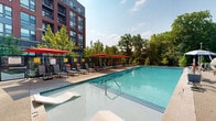 This two-level outdoor amenity space is designed to deliver a resort-style experience within an urban residential setting. At its centerpiece sits a generously sized swimming pool with crystal-clear blue water, flanked by a broad sun deck outfitted with rows of lounge chairs shaded by bold red market umbrellas. Adjacent to the main pool, a shallow tanning ledge with built-in loungers offers a relaxed spot to unwind at water's edge. A covered indoor lounge area with floor-to-ceiling glass walls and vibrant orange chaise lounges provides a sheltered vantage point directly overlooking the pool, bathed in natural light throughout the day. Landscaped terraces with flowering perennials, ornamental grasses, and young deciduous trees add greenery and texture throughout the space, softened further by tiered concrete staircases with dark metal railings connecting the two levels. The upper level features glass-panel railings along a wide walkway, offering open views of the tree-lined boulevard and surrounding neighborhood. The building's striking red brick and large-format window façade frames the entire amenity area, lending an architectural backdrop that is both contemporary and refined. Secure gated access points ensure privacy throughout the space.