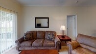 This single-floor residence offers a well-organized layout connecting several functional living spaces. The living room serves as the central gathering area, finished with plush carpeting, crown molding, a ceiling fan with light fixture, and a sliding glass door that brings in natural light and provides views of the outdoors. The space accommodates comfortable seating arrangements alongside a television setup.

The galley-style kitchen features white cabinetry lining both walls, white appliances including a refrigerator, electric range, and dishwasher, complemented by tile flooring and a double-basin sink. It opens directly into a dedicated dining area, which is finished with crown molding and provides a natural transition between the kitchen and living spaces. A passthrough also connects the kitchen corridor to the main hallway.

The primary bedroom is generously sized with carpeted floors, a ceiling fan, a window for natural light, and direct access to a bathroom equipped with a tub-shower combination and tile flooring. A second bedroom mirrors similar finishes, including carpeting and a ceiling fan. A second full bathroom also features a tub-shower combination. In-unit washer and dryer connections are visible within the layout, adding practical convenience to this thoughtfully arranged home.