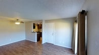 This single-floor space offers a clean, functional layout designed for comfortable everyday living. The main living area features warm wood-look plank flooring that extends throughout, complementing the neutral grey walls and white trim for a modern, cohesive aesthetic. A textured popcorn ceiling adds character overhead, while a curtained window along one wall allows for natural light and privacy control. A classic six-panel white entry door serves as a prominent focal point, providing a welcoming entryway directly into the living space. A half-wall pass-through connects the living area to the kitchen, creating an open, communicative flow between the two spaces without fully removing the sense of separation. The kitchen features dark cabinetry with recessed lighting above, offering a rich contrast to the lighter tones found throughout the rest of the space. The design emphasizes practicality and an open feel, with clearly defined zones that work together harmoniously. The neutral color palette and durable flooring make this a versatile space that can be easily personalized to suit a variety of interior styles.