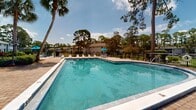 This outdoor recreational area centers around a generously sized swimming pool with clear, blue-tinted water, designed for both leisure and lap swimming. The pool is framed by a wide brick paver deck that provides ample space for sunbathing and socializing. A collection of lounge chairs and outdoor dining sets with blue umbrellas are arranged across the deck, offering shaded seating options throughout the day.

A distinctive two-story gazebo-style clubhouse building serves as a focal point of the area. Painted in a light blue exterior, it features a covered upper observation deck and is surrounded by vibrant tropical landscaping, including red-leafed plants, palms, and tall pine trees that provide natural shade and a lush backdrop. An American flag and decorative banners are displayed prominently on flagpoles nearby.

The entire pool area is enclosed by a black wrought-iron fence with a gated entrance, and is designated as a smoke-free zone. A separate hot tub or spa is also visible adjacent to the main pool. The surrounding landscape is rich with mature pine and palm trees, lending a Florida-inspired, park-like atmosphere to the space. The overall setting is clean, well-maintained, and designed to maximize outdoor enjoyment in a natural, tree-shaded environment.