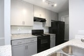 This single-floor residence is designed with a practical, open-concept layout that connects the living and kitchen areas seamlessly. The kitchen is configured as a galley-style space with a peninsula that opens toward the main living area. It features white shaker-style cabinetry, granite-look countertops, a subway tile backsplash, black appliances including a range and refrigerator, and track lighting overhead. Dark wood-look flooring runs throughout the kitchen and hallway, providing a sharp contrast to the light gray walls.

The living area is generously proportioned, finished with neutral carpet and illuminated by natural light from large windows fitted with blinds. A pendant light anchors the dining zone near the kitchen peninsula, and a textured ceiling adds subtle visual interest throughout the common areas.

The bedrooms are carpeted and feature neutral gray walls, with multiple windows in the primary bedroom allowing for ample natural light. The bathroom includes a full tub-shower combination with white tile surround, chrome fixtures, and a granite-topped vanity with gray cabinetry. A closet with louvered doors is conveniently positioned nearby. Overall, the unit presents a clean, updated, and cohesive aesthetic.