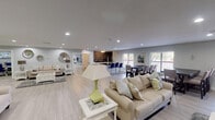 This single-floor space is designed as a well-appointed communal gathering area, combining multiple functional zones within an open, flowing layout. Light wood-style flooring runs continuously throughout, contributing to a clean, cohesive aesthetic that unifies the various areas.

The main lounge area is generously proportioned, featuring multiple seating arrangements with neutral-toned sofas, white side tables, and accent lamps. A soft green accent wall with a built-in display niche adds a distinctive architectural detail, while recessed lighting and a wall-mounted television enhance the room's functionality. Decorative metallic wall art provides additional visual interest in a secondary lounge area, which connects seamlessly to the rest of the space.

A dedicated dining area accommodates group seating around a large dark wood table with upholstered chairs and a bench, positioned near windows fitted with blinds that allow natural light to filter in. Adjacent to this is a kitchenette featuring white cabinetry, dark countertops, stainless steel appliances, and a striking stacked-stone accent wall, adding texture and warmth. A separate windowed office or workroom is also accessible via glass-paneled doors, completing this versatile, multi-purpose space.