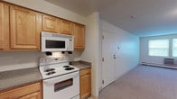 This single-floor 1-bedroom, 1-bathroom unit offers a practical and well-organized layout. The kitchen is equipped with warm-toned wood cabinetry providing generous storage, granite-style laminate countertops, and a full suite of white appliances including an electric range, over-the-range microwave, dishwasher, and refrigerator. Light tile flooring runs throughout the kitchen, which connects seamlessly to the rest of the unit. The living area is spacious and open, featuring neutral wall-to-wall carpeting, baseboard heating, and a wall-mounted air conditioning unit. Windows allow natural light to filter in, offering views of surrounding greenery. A secondary door with a glass panel provides additional access and further brightens the space. The bedroom is a generous, carpeted room with baseboard heating and ample wall space for flexible furniture arrangement. The full bathroom features white cabinetry with an integrated vanity, a mirrored medicine cabinet, a tiled tub and shower combination, and tile flooring, all accented with chrome fixtures. Overall, the unit presents a clean, functional design with a neutral palette throughout.