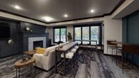 Located at 486–498 Wacouta St in Saint Paul, Minnesota, this single-floor community amenity space is thoughtfully designed to blend comfort, functionality, and modern style. The lounge is organized into distinct yet connected zones, each serving a different social purpose.

The living area anchors the space with a plush sofa accented by mustard throw pillows, a rustic wooden coffee table, and a tiled fireplace framed by a bold teal accent wall — creating a warm, inviting atmosphere. Large black-trimmed windows line one side of the room, filling the interior with natural light and offering views of the surrounding greenery and neighborhood streetscape.

A central high-top communal table with industrial-style bar stools serves as a flexible workspace or casual gathering spot. Adjacent to this is a dedicated wet bar area, featuring rich dark wood cabinetry with gold hardware, a sleek black countertop, a built-in wine refrigerator, and a striking floor-to-ceiling striped stone accent wall. Pendant lights hang above the bar, adding warmth and a curated aesthetic.

The game area rounds out the space with a shuffleboard table set against the teal wall, ideal for entertainment and social play. Textured area rugs throughout tie the zones together, while crown molding and recessed lighting add a refined, polished finish to the overall design.