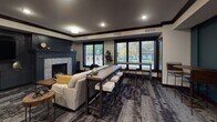 Located at 486–498 Wacouta St in Saint Paul, Minnesota, this single-floor community amenity space is thoughtfully designed to blend comfort, functionality, and modern style. The lounge is organized into distinct yet connected zones, each serving a different social purpose.

The living area anchors the space with a plush sofa accented by mustard throw pillows, a rustic wooden coffee table, and a tiled fireplace framed by a bold teal accent wall — creating a warm, inviting atmosphere. Large black-trimmed windows line one side of the room, filling the interior with natural light and offering views of the surrounding greenery and neighborhood streetscape.

A central high-top communal table with industrial-style bar stools serves as a flexible workspace or casual gathering spot. Adjacent to this is a dedicated wet bar area, featuring rich dark wood cabinetry with gold hardware, a sleek black countertop, a built-in wine refrigerator, and a striking floor-to-ceiling striped stone accent wall. Pendant lights hang above the bar, adding warmth and a curated aesthetic.

The game area rounds out the space with a shuffleboard table set against the teal wall, ideal for entertainment and social play. Textured area rugs throughout tie the zones together, while crown molding and recessed lighting add a refined, polished finish to the overall design.