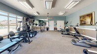 The fitness center at 345 Stone Park Blvd in Pike Road, Alabama, offers a well-appointed single-floor exercise space designed to support a comprehensive workout routine. The room features soft blue-gray walls accented by white wainscoting, giving the space a clean and inviting atmosphere. Recessed and overhead fluorescent lighting ensures the entire area is well illuminated throughout the day.

The cardio section is equipped with multiple treadmills, elliptical trainers, a stationary bike, and a recumbent bike, all arranged to allow comfortable spacing between machines. A wall-mounted flat-screen television is positioned for easy viewing during workouts. The strength training area includes a multi-station cable and weight machine, a free-weight rack with dumbbells, and a weight bench, providing options for full-body resistance training.

Large windows along one wall allow natural light to fill the space and offer views of the outdoor surroundings, including what appears to be a patio or pool area. A large mirror reflects additional light and helps users monitor their form. Neutral-toned carpet tile covers the floor throughout, and a ceiling fan adds to the comfort of the space. Decorative framed artwork adds a polished finishing touch to the overall environment.
