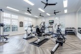 Located at 3812 NW Barry Rd in Kansas City, MO, this fitness center offers a thoughtfully designed single-floor space dedicated to comprehensive exercise and wellness. The interior is finished with light-toned herringbone-patterned flooring throughout, lending a clean, modern aesthetic that complements the bright white walls and crisp trim.

Natural light pours in through multiple large windows positioned along the perimeter walls, providing views of the surrounding outdoor greenery and creating an open, airy atmosphere. The ceiling features multiple black ceiling fans alongside recessed and panel lighting, ensuring consistent airflow and visibility across the entire space.

The cardio area is well-stocked with treadmills, ellipticals, stationary bikes, a rowing machine, and a stair climber. A wall-mounted flat-screen television is positioned for easy viewing during workouts. On one side of the room, a full-length mirror wall visually expands the space while providing functional feedback for form and technique.

The strength training section includes a multi-functional cable machine, a dedicated free weight area with tiered dumbbell racks, and a weight bench. An additional support area with storage and utility fixtures is accessible from the main floor. Overall, the space delivers a professional, well-maintained environment suited for a wide range of fitness activities.