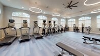 Located in Bradenton, FL, this well-appointed fitness center offers a comprehensive workout environment designed for both cardio and strength training. The single-floor facility is characterized by its clean, modern aesthetic, featuring light wood-style flooring, neutral gray and warm yellow accent walls, and a series of distinctive circular ring pendant lights that illuminate the space with a contemporary glow. Large arched windows line the perimeter walls, flooding the interior with abundant natural light and providing views of the surrounding greenery outdoors. Overhead ceiling fans contribute to a comfortable training environment throughout the facility. The cardio zone is extensively equipped with multiple treadmills, elliptical trainers, stair steppers, and Peloton-style stationary bikes, all featuring integrated digital displays. The strength training area houses a full rack of dumbbells, adjustable weight benches, a cable machine system, and a multi-function strength training tower, all reflected in a large floor-to-ceiling mirror wall that visually expands the space. Glass-paneled doors provide access to outdoor areas, enhancing the sense of openness. The facility's spacious layout ensures ample room between equipment, creating an organized and functional workout environment suited to a wide range of fitness routines.
