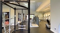 This fitness center offers a comprehensive workout environment thoughtfully organized across a single floor. The space is divided into functional zones, beginning with a strength training area anchored by a multi-use power rack, free weights, and a stability ball, providing ample room for resistance and functional training exercises. Adjacent to this zone, a selection of weight machines offers targeted muscle group training for users of all fitness levels. The cardio section lines one wall with a row of treadmills and additional aerobic equipment, all positioned beneath linear ceiling-mounted lighting that ensures consistent, bright illumination throughout the space. The layout is open and spacious, with high vaulted ceilings adding to the sense of airiness. Light-colored walls and large windows allow natural light to filter in, creating an inviting and energizing atmosphere. The dark rubberized flooring throughout provides both durability and safety, ideal for high-impact activity. A ceiling fan aids in air circulation, contributing to a comfortable workout environment. The overall design is clean and modern, with a neutral color palette that keeps the focus on functionality and performance.