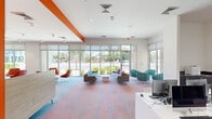 Located at 1100 East Apache Boulevard in Tempe, Arizona, this single-floor communal space offers a generously sized, open-plan layout designed for both collaboration and relaxation. The interior is immediately distinguished by its bold, energetic color palette — vivid orange accent walls adorned with geometric white relief detailing serve as striking focal points throughout the space.

Floor-to-ceiling windows line multiple walls, flooding the interior with abundant natural light and offering unobstructed views of the surrounding Tempe streetscape and landscaping. A vibrant multicolored carpet in coral and blue tones anchors the space and ties together its diverse functional zones.

The lounge area features a thoughtful mix of seating arrangements, including modular sofas with coral accent pillows, teal pod chairs, and casual groupings centered around round white coffee tables — ideal for informal gatherings. A dedicated computer workstation area lines one wall with desktop terminals and sleek white countertops. Bright yellow chairs accompany white rectangular tables in a collaborative study or dining zone, while wood-paneled booth seating with red upholstery and tabletops provides additional semi-private options.

A glass-walled interior room with colorful hanging pendant lights adds a dynamic visual layer, functioning as a private meeting or activity space. The overall ambiance is modern, youthful, and highly functional, catering to an active, community-oriented environment.